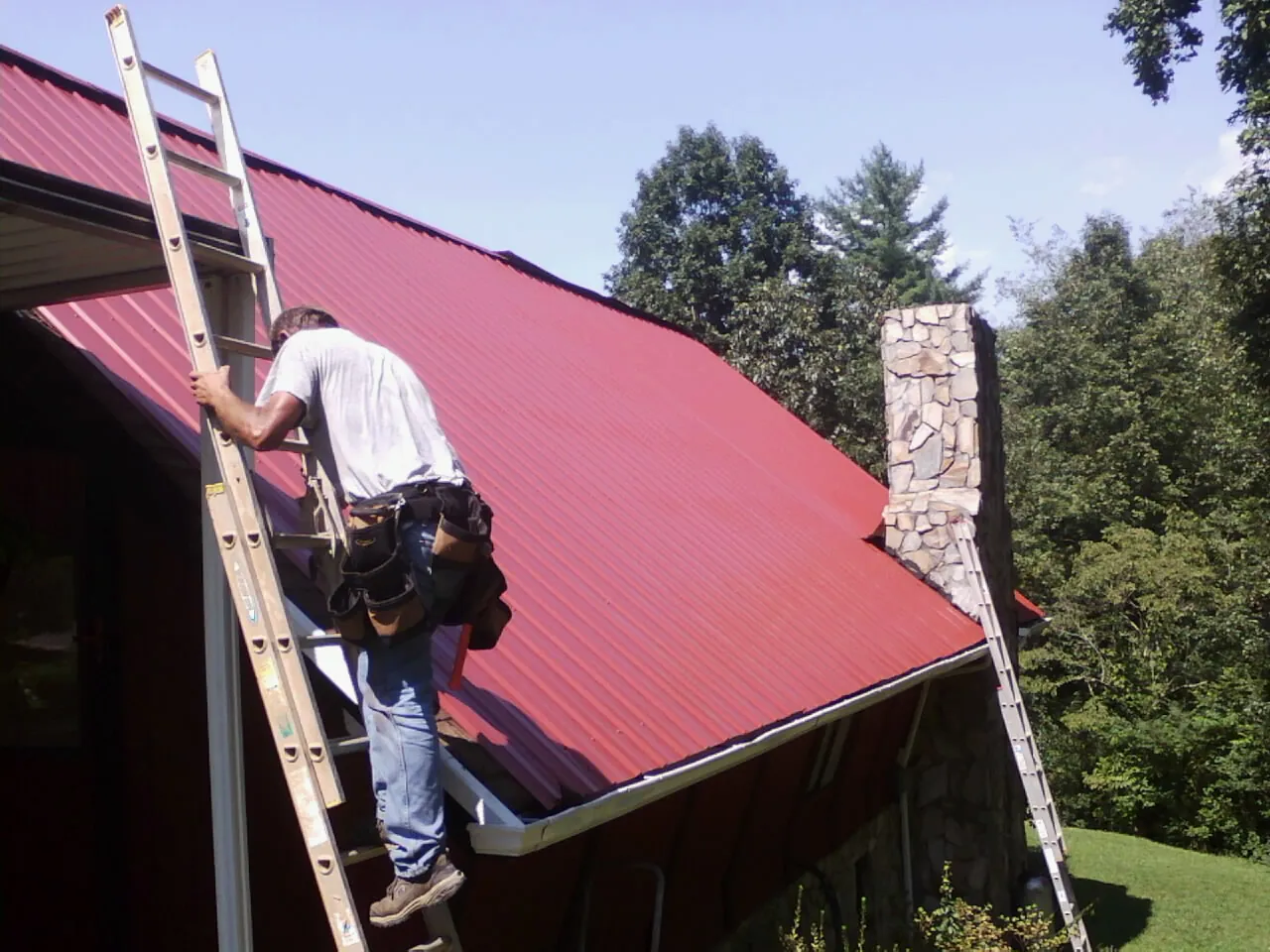 Roofer on a ladder beside a red metal roof and stone chimney — Soffit & Fascia Repair by Roofing Brothers in Far Rockaway